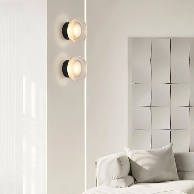 "Two modern, minimalist wall lamps with circular glass diffusers and sleek black frames mounted on a white wall in a living room, flanking a textured art piece above a white sofa."