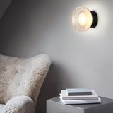 "A modern, minimalist wall lamp with a circular glass diffuser and a sleek black frame mounted on a gray wall in a living room, providing ambient lighting next to a cozy armchair and a side table with books."