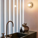 "Two modern, minimalist wall lamps with circular glass diffusers and sleek brass frames mounted on a light pink wall in a contemporary bathroom, providing ambient lighting above a dark marble sink and vanity."