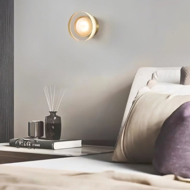 "A modern, minimalist wall lamp with a circular glass diffuser and a sleek brass frame mounted on a gray wall in a contemporary bedroom, providing ambient lighting above a nightstand with decorative objects."