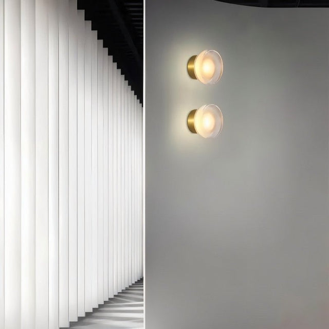"Two modern, minimalist wall lamps with circular glass diffusers and sleek brass frames mounted on a gray wall in a hallway, providing ambient lighting and a design element next to a textured white wall."