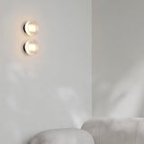 "Two modern, minimalist wall lamps with circular glass diffusers and sleek brass frames mounted on a white wall in a living room, providing ambient lighting above a white sofa."