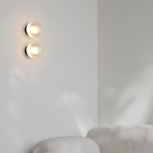 "Two modern, minimalist wall lamps with circular glass diffusers and sleek brass frames mounted on a white wall in a living room, providing ambient lighting above a white sofa."