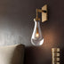 Pulcher Glass Tears wall sconce with gold finish and clear glass shade mounted on dark textured wall in interior setting.