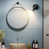 "A black wall lamp with a round base and a cylindrical shade, shown lit up on a light blue wall above a round mirror, next to a sink with soap dispensers and a vase with flowers."