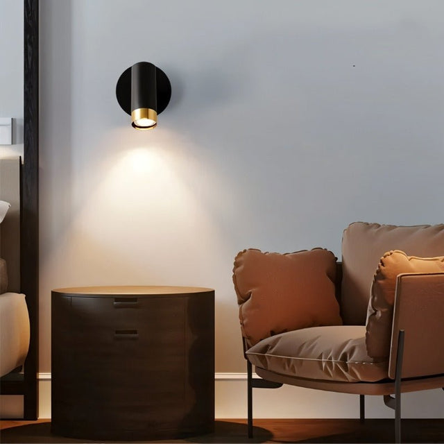 "A black wall lamp with a round base and a cylindrical shade, shown lit up on a light gray wall above a brown nightstand, next to a brown armchair with pillows."
