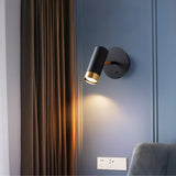 "A black wall lamp with a round base and a cylindrical shade, shown lit up on a light blue wall next to a brown curtain."