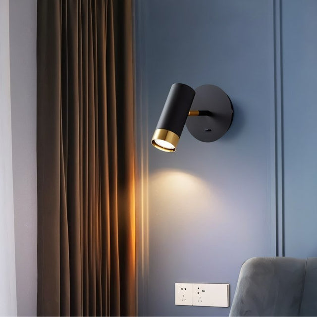 "A black wall lamp with a round base and a cylindrical shade, shown lit up on a light blue wall next to a brown curtain."