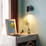 "A black wall lamp with a round base and a cylindrical shade, shown lit up on a light blue wall above a white vanity with a mirror, a vase with flowers, and makeup products."