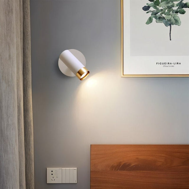 "A white wall lamp with a round base and a cylindrical shade, shown lit up on a light gray wall next to a framed artwork of a plant, above a bed with a wooden headboard."