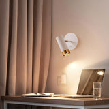 "A white wall lamp with a round base and a cylindrical shade, shown lit up on a light gray wall above a desk with a laptop, a book, and a glass of water."