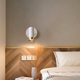 "A white wall lamp with a round base and a cylindrical shade, shown lit up on a light gray wall above a wooden headboard with a chevron pattern, next to a nightstand with a vase and a book."