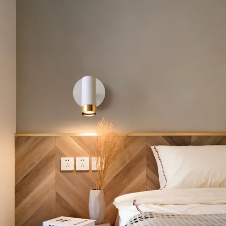 "A white wall lamp with a round base and a cylindrical shade, shown lit up on a light gray wall above a wooden headboard with a chevron pattern, next to a nightstand with a vase and a book."