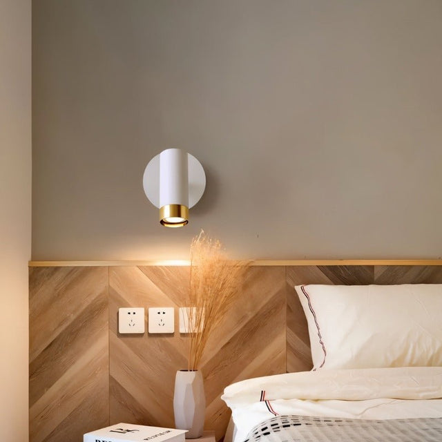 "A white wall lamp with a round base and a cylindrical shade, shown lit up on a light gray wall above a wooden headboard with a chevron pattern, next to a nightstand with a vase and a book."
