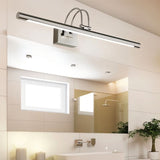 "A silver wall lamp with a linear LED bar mounted above a bathroom mirror, showing the full bathroom setting with tiles, window, and plants."