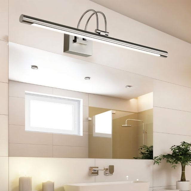 "A silver wall lamp with a linear LED bar mounted above a bathroom mirror, showing the full bathroom setting with tiles, window, and plants."