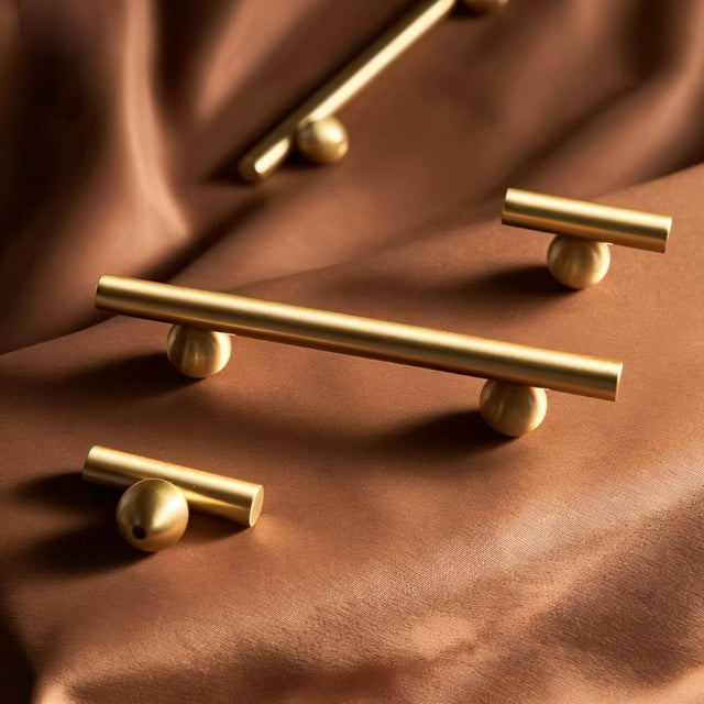 "Gold brass knobs and pull bars with cylindrical design, arranged on a brown satin fabric."