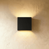 Brass square wall sconce with warm glow mounted on mauve wall above wooden chair in lifestyle setting.
