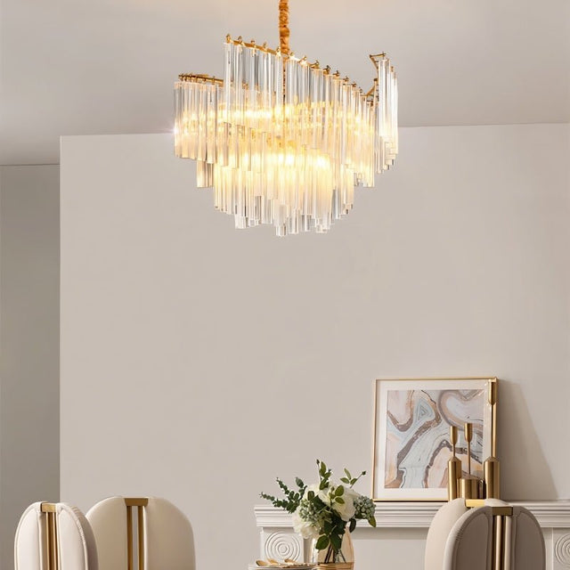 "A tiered chandelier with clear crystal rods and a brass frame, hanging above a dining table in a well-lit room with artwork."