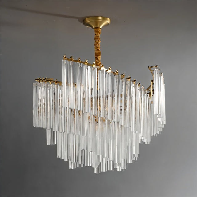 "A tiered chandelier with clear crystal rods and a brass frame, suspended against a plain grey backdrop."