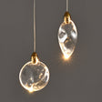 "Two uniquely shaped crystal pendant lights, one round and one oval, with gold caps, hang against a grey background, showcasing their elegant design and warm light."