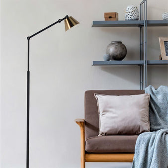 "A modern floor lamp with a black adjustable arm and gold shade, providing focused lighting in a cozy reading nook with a comfortable armchair and shelving."