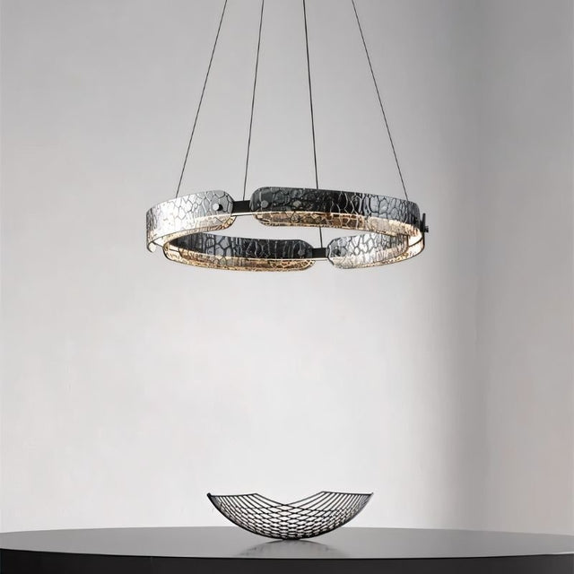 "A modern, circular chandelier with a segmented ring design and textured pattern, made of iron and glass, suspended by thin cables above a dining table."