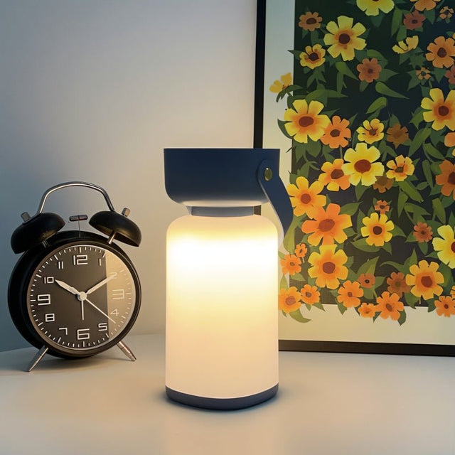 "A blue and white cylindrical table lamp with a handle, lit up with a soft glow, sitting on a table next to a black alarm clock and a framed floral artwork."