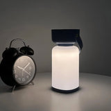 "A black and white cylindrical table lamp with a handle, lit up with a soft glow, sitting on a table next to a black alarm clock."