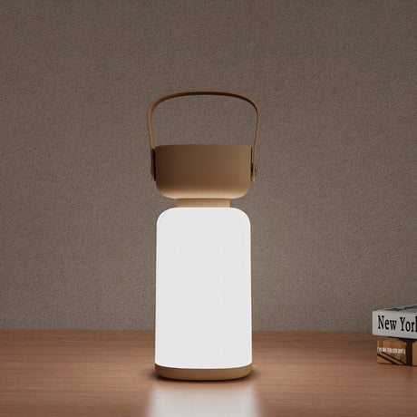 "A beige and white cylindrical table lamp with a handle, lit up with a soft glow, sitting on a wooden table with books."