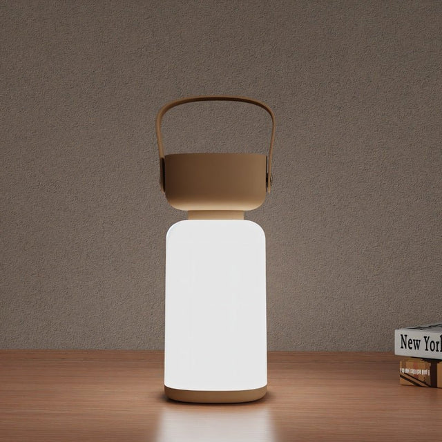 "A beige and white cylindrical table lamp with a handle, lit up with a soft glow, sitting on a wooden table with books."