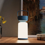 "A blue and white cylindrical table lamp with a handle, lit up with a soft glow, sitting on a wooden table with books and a framed painting."