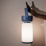 "A blue and white cylindrical table lamp with a handle, lit up with a soft glow, hanging from a branch against a light wall."