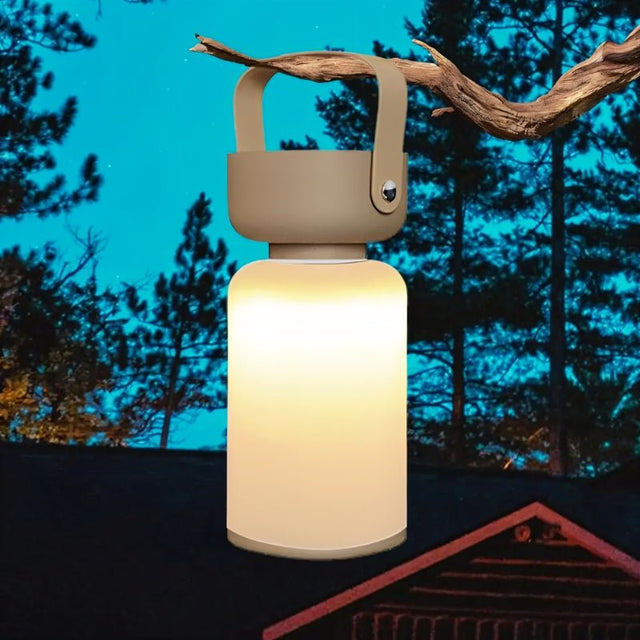 "A beige and white cylindrical table lamp with a handle, lit up with a bright glow, hanging from a tree branch outdoors at dusk."