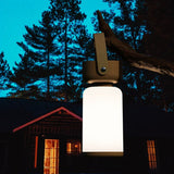 "A beige and white cylindrical table lamp with a handle, hanging from a tree branch outdoors at dusk."