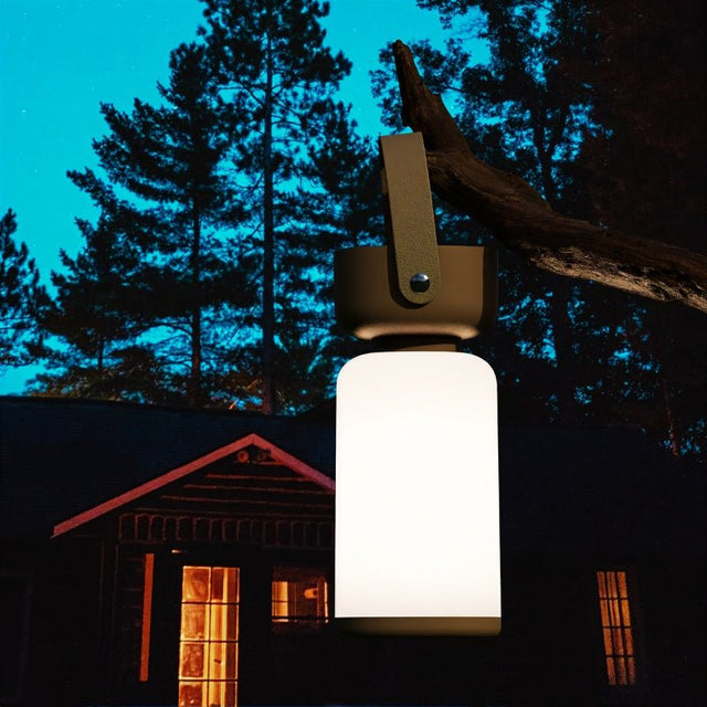 "A beige and white cylindrical table lamp with a handle, hanging from a tree branch outdoors at dusk."