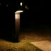 Modern black outdoor bollard lamp with horizontal slats, illuminated at night in a residential garden setting.