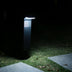Modern black outdoor bollard lamp with horizontal slats emitting bright white light, installed in grass at night.