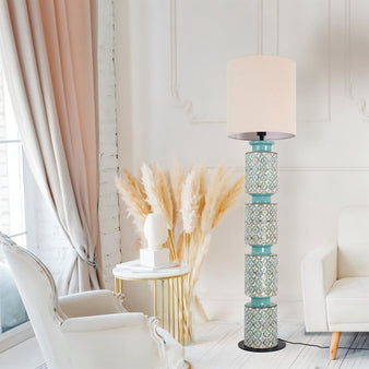 "Floor lamp with a stacked ceramic base in aqua blue and orange geometric patterns, paired with a neutral fabric shade, placed next to a white armchair and decorative pampas grass in a living room."