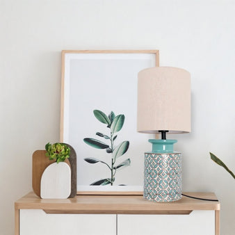 "Table lamp with a patterned ceramic base in aqua blue and orange, paired with a neutral fabric shade, on a wooden dresser with a framed picture."