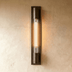 Vertical brass wall lamp with cylindrical glass shade on dark gray background.