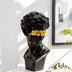 Black resin bust sculpture with gold-toned blindfold displayed on white pedestal in modern interior setting.