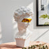 White plaster bust sculpture with gold metallic eye mask, displayed on wooden surface with green plant foliage.