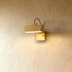 Gold brass modern wall sconce with curved arm mounted on white textured wall, countryside landscape visible in background.