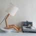 Wooden articulated desk lamp with white shade on white pedestal base, displayed with books and decorative items.