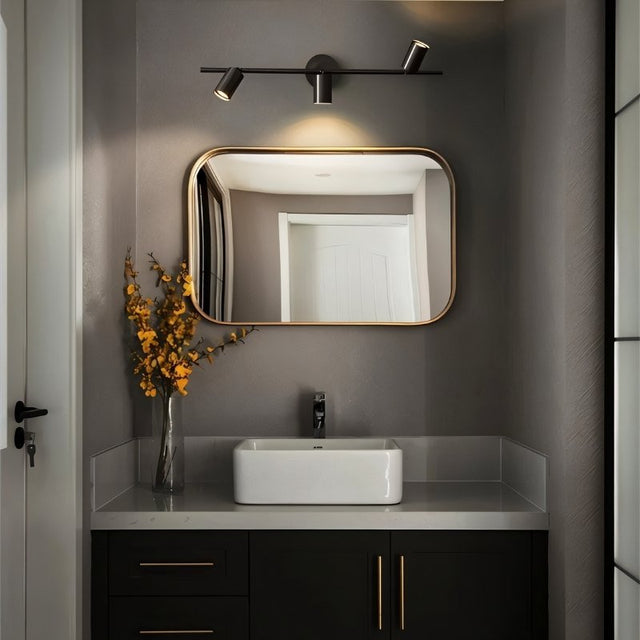 "A modern black bathroom vanity light with three adjustable heads mounted on a circular base above a rounded rectangular mirror."