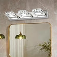 "A modern crystal and stainless steel wall lamp mounted on a textured wall, illuminating a bathroom featuring a gold-framed mirror and hanging pendant light."