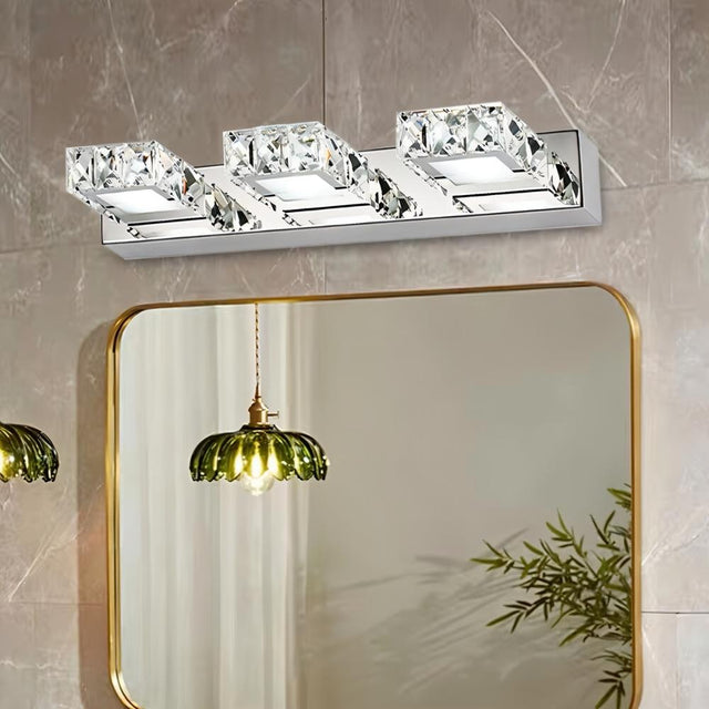 "A modern crystal and stainless steel wall lamp mounted on a textured wall, illuminating a bathroom featuring a gold-framed mirror and hanging pendant light."