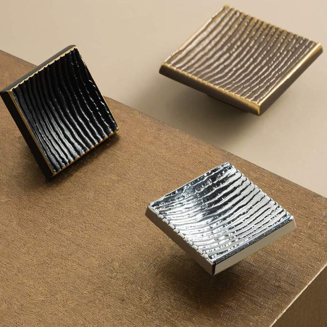 "Studio shot of modern square brass cabinet knobs with a textured, ridged surface in black, bronze, and silver finishes."