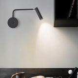 "A black wall lamp with a pivoting arm and cylindrical head, installed above a desk with a book titled 'MEMOIRS OF A GEEZER', a mug, and glasses."